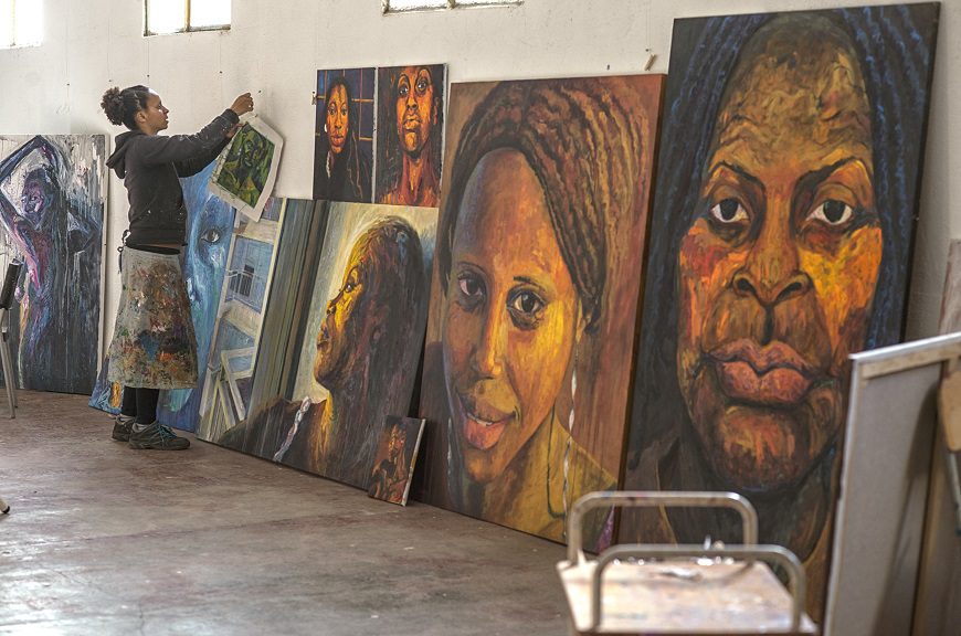 La artista Bianca al fondo colocando un cuadro en una pared llena de grandes de sus obras, pinturas, que reflejan a mujeres negras