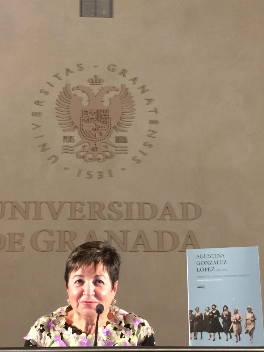 Una mujer de hombros para arriba, detrás de una mesa junto al libro que presenta. De fondo, el logo de la Universidad de Granada